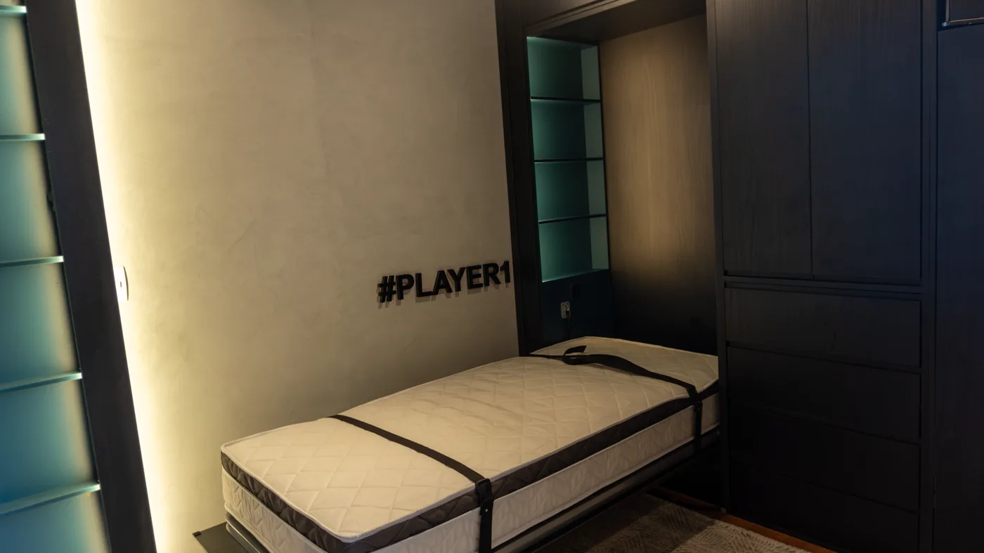 Cama aberta com colchão no studio, telão e setup gamer ao lado